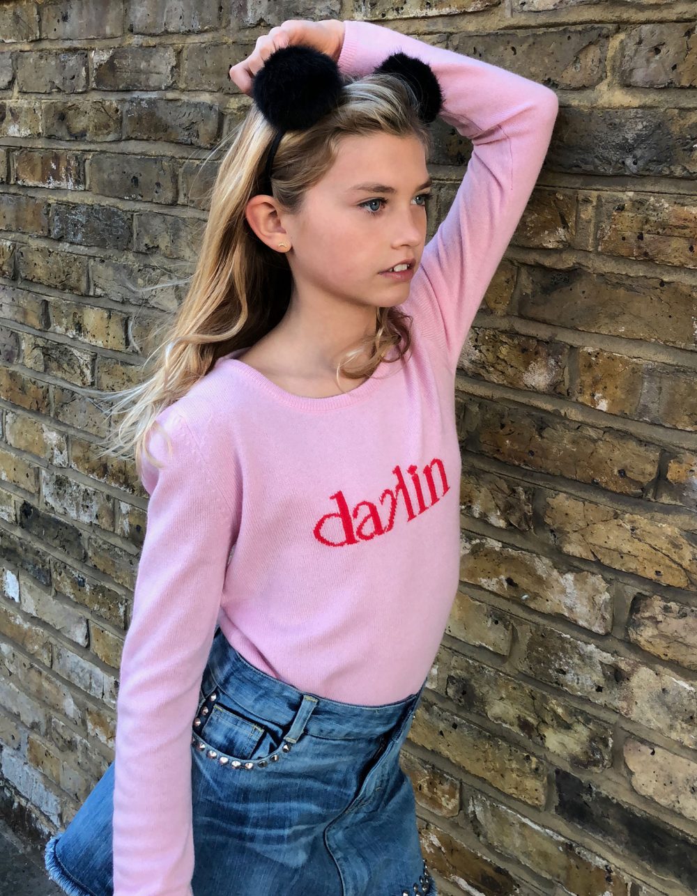 A girl leaning against a brick wall, wearing a malin darlin Little Darlin pink kids cashmere jumper.