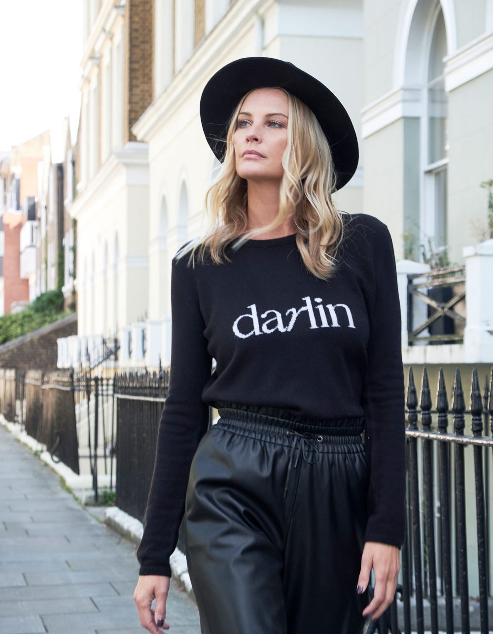 Model on a residential street wearing a hat and black cashmere jumper with the word darlin on the front.